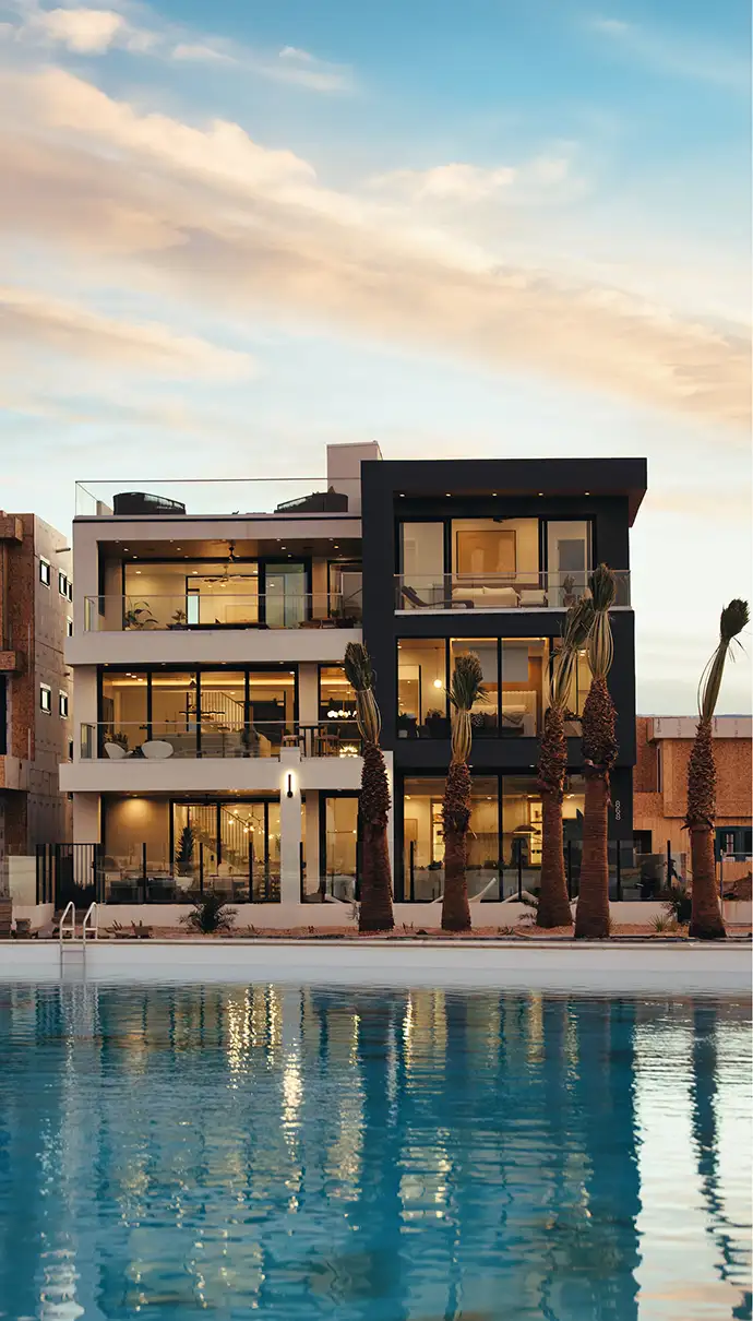Modern three-story home with lit windows and palm trees beside a lagoon at dusk