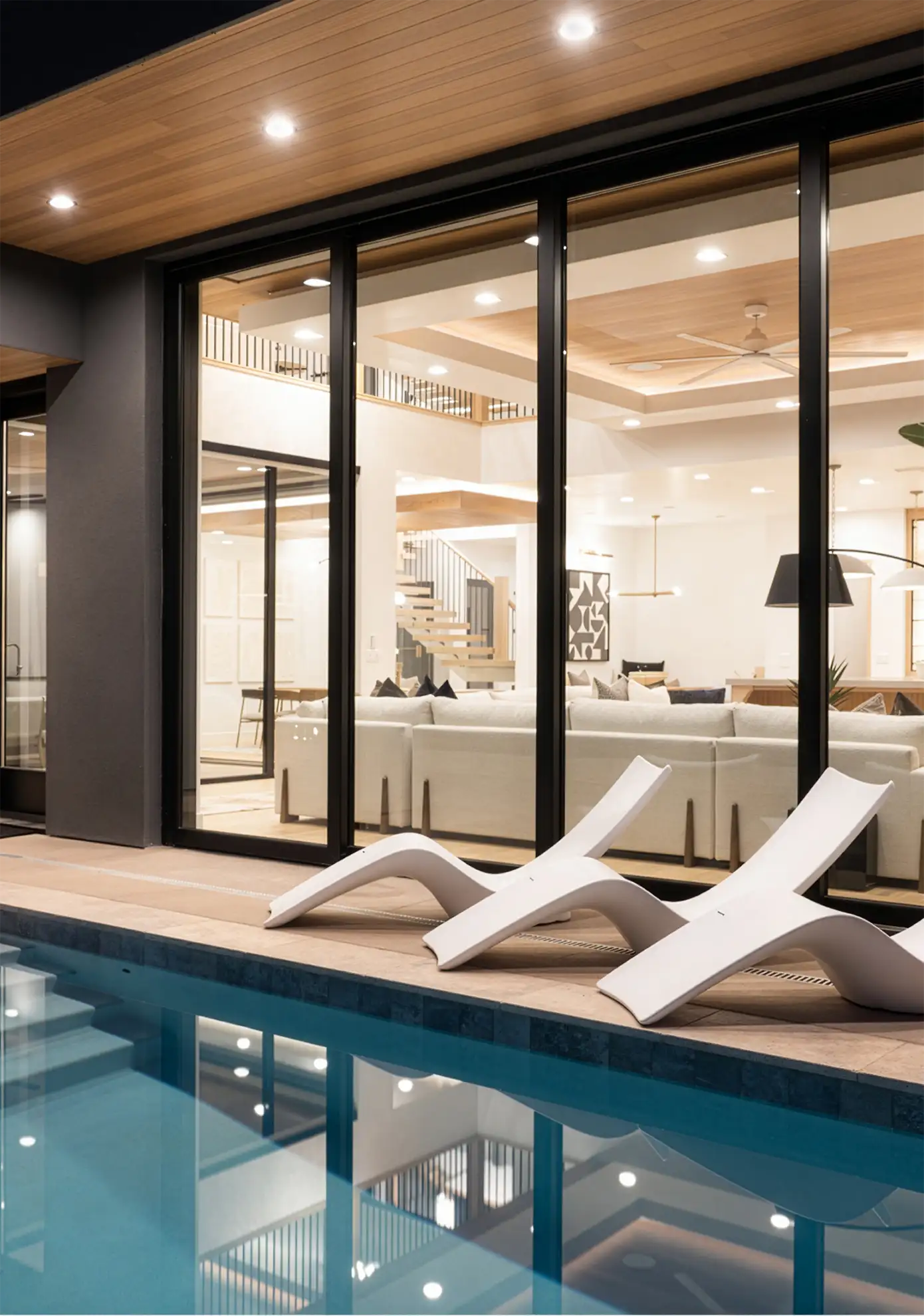 Backyard pool and white lounge chairs beside glass doors into a lit living room at night