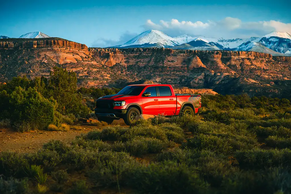 Red pickup truck parked among desert brush below red rock cliffs and snowy mountains