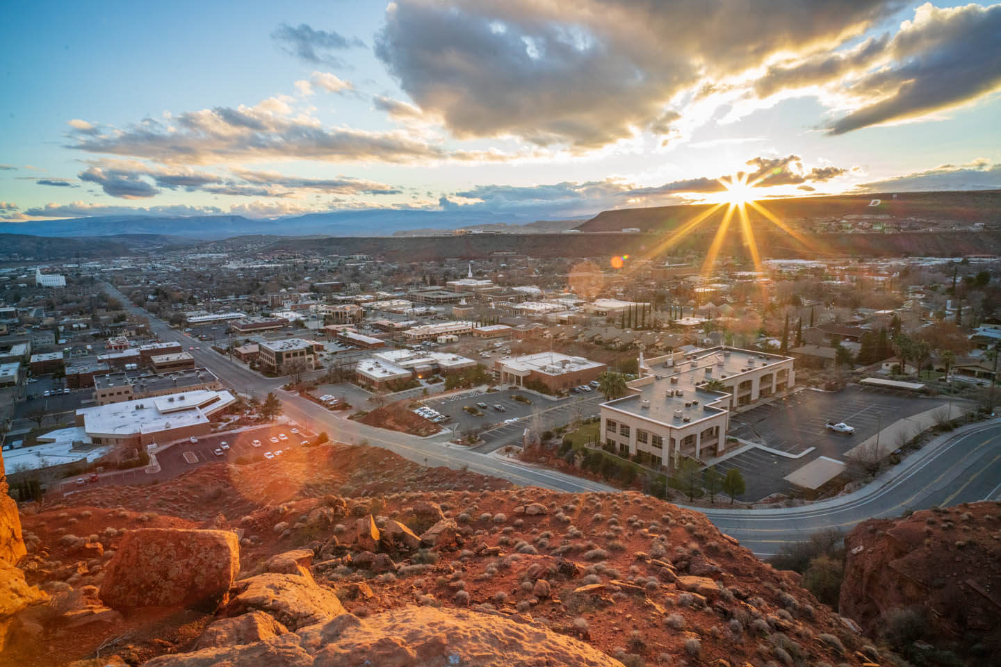 Sunset over a small city with red rocky foreground and clouded sky.