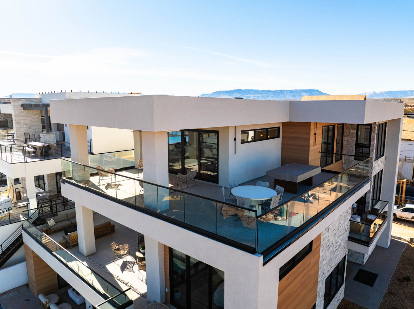 Modern two-story house with large glass balconies, outdoor seating, and mountain views under a clear blue sky.