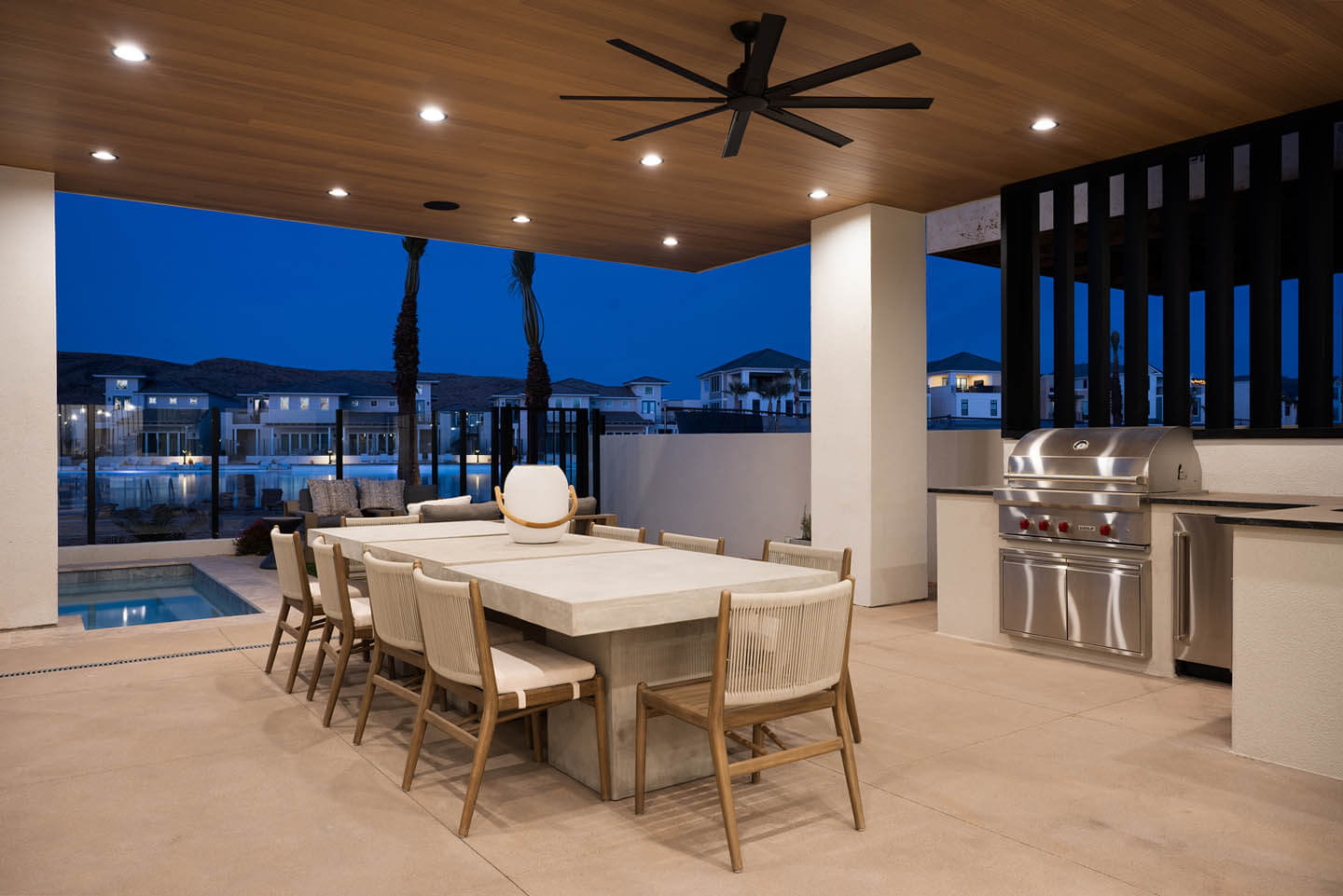 Modern outdoor patio with a large rectangular dining table surrounded by eight chairs, ceiling fan, built-in stainless steel grill, and a pool with a view of nearby houses at dusk.