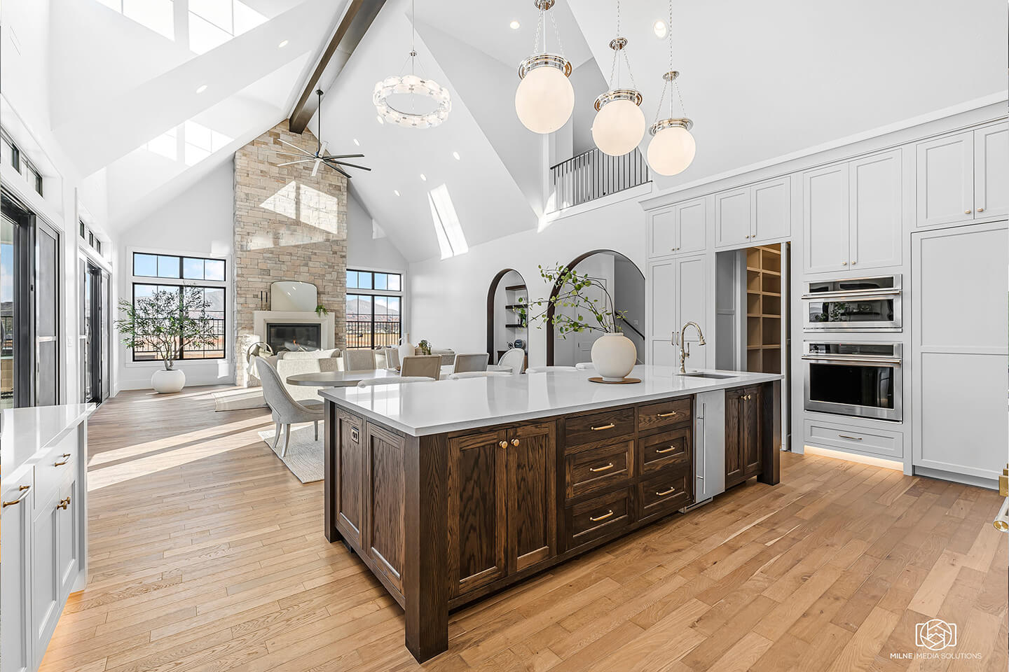 Bright open-concept kitchen and living area with vaulted ceilings, large island with dark wood cabinets and white countertop, pendant lights, and a stone fireplace.