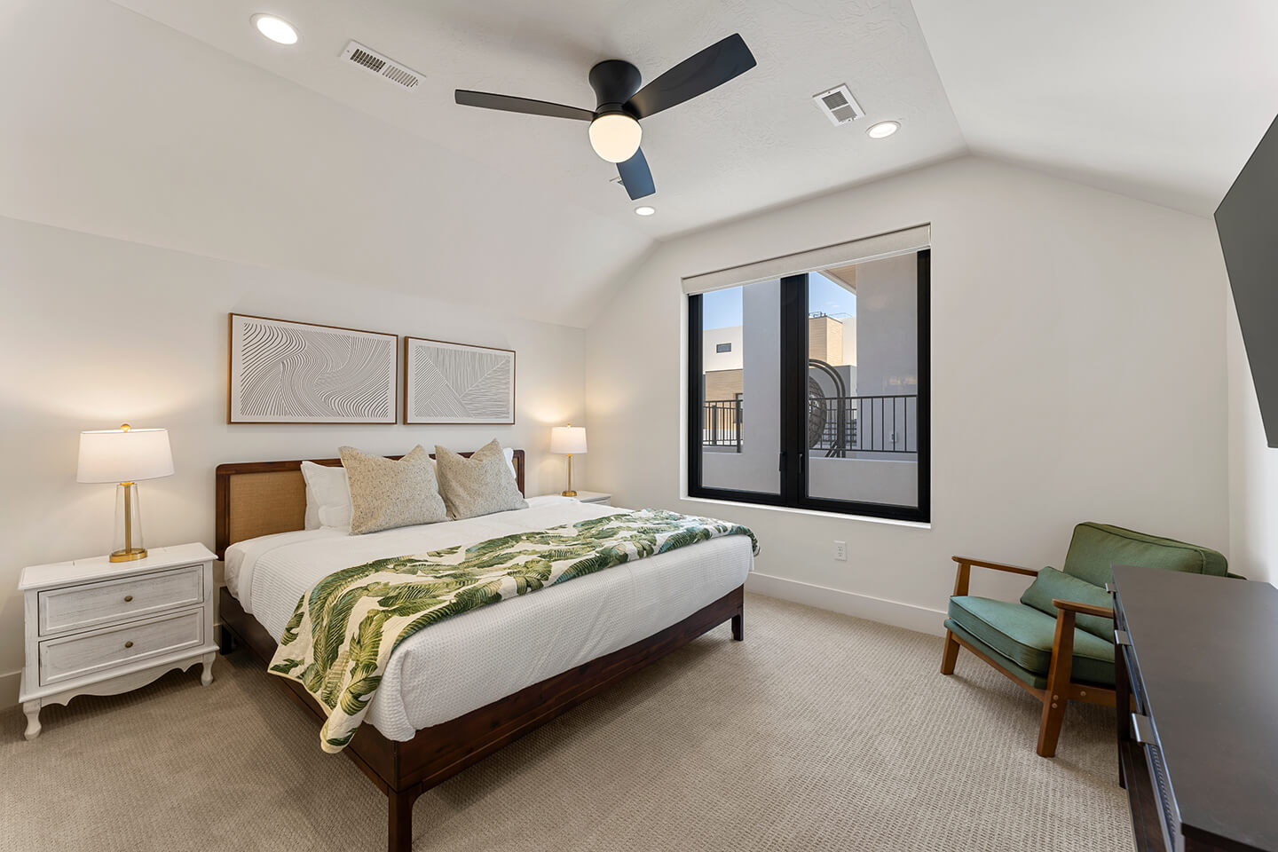 Modern bedroom with a wooden bed, white bedding, green leaf-patterned blanket, two nightstands with lamps, an armchair, and a window with a view outside.