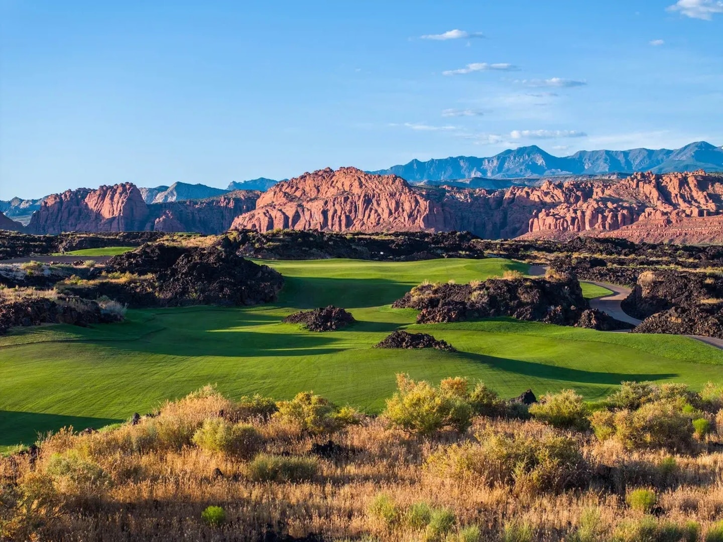 Lush green fairways of the Black Desert golf course winding through dramatic black lava rock formations with red sandstone cliffs in the background
