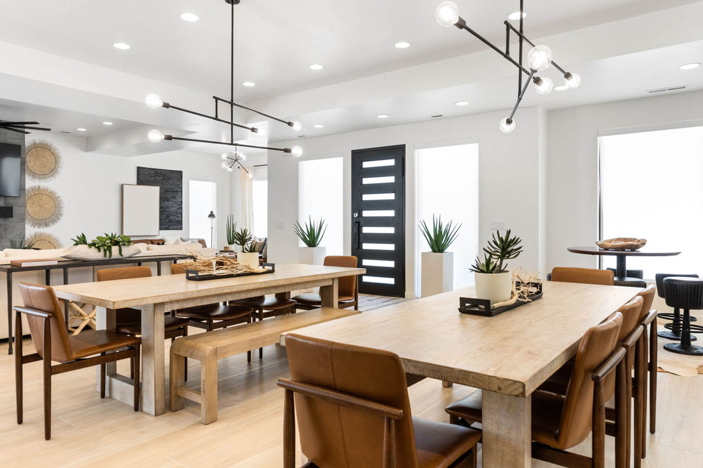 Expansive dining area with two large natural wood dining tables paired with camel leather chairs, illuminated by a modern black globe chandelier spanning the length of the room. The open floor plan connects to the living room with its stone fireplace and t
