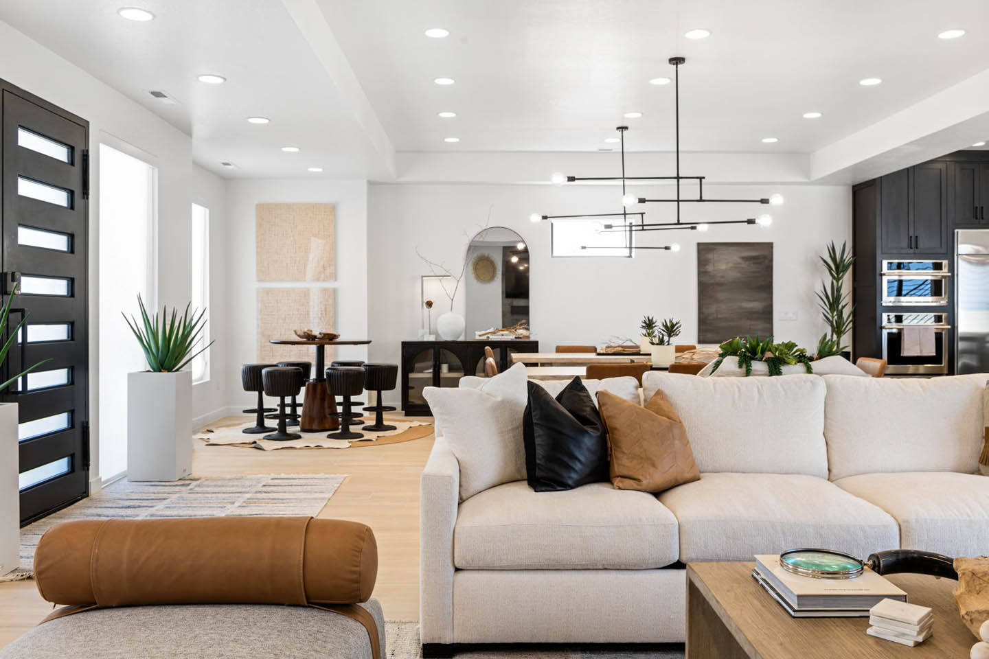 Open-concept great room view looking from the living area toward the entry and kitchen, showcasing a cream sectional sofa with black and tan accent pillows, a modern black linear chandelier overhead, light hardwood floors, and a modern black front door wit