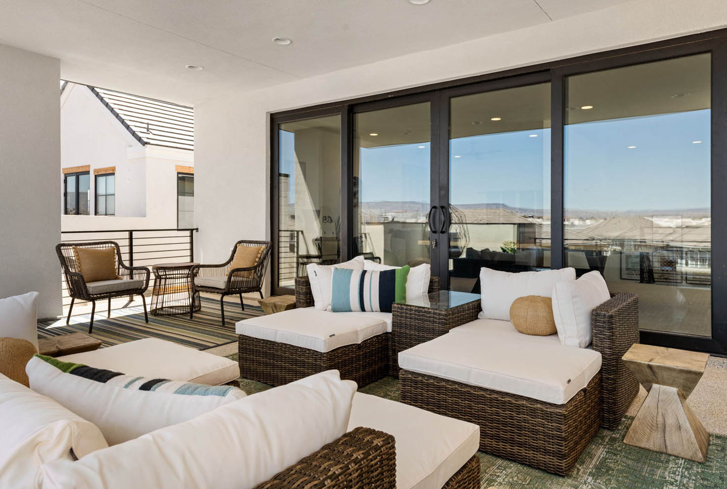 Covered upper-level patio furnished with wicker lounge chairs and ottomans with white cushions, accented by striped and colorful throw pillows, alongside a pair of rattan bistro chairs. Large sliding glass doors connect to the interior, while the open side