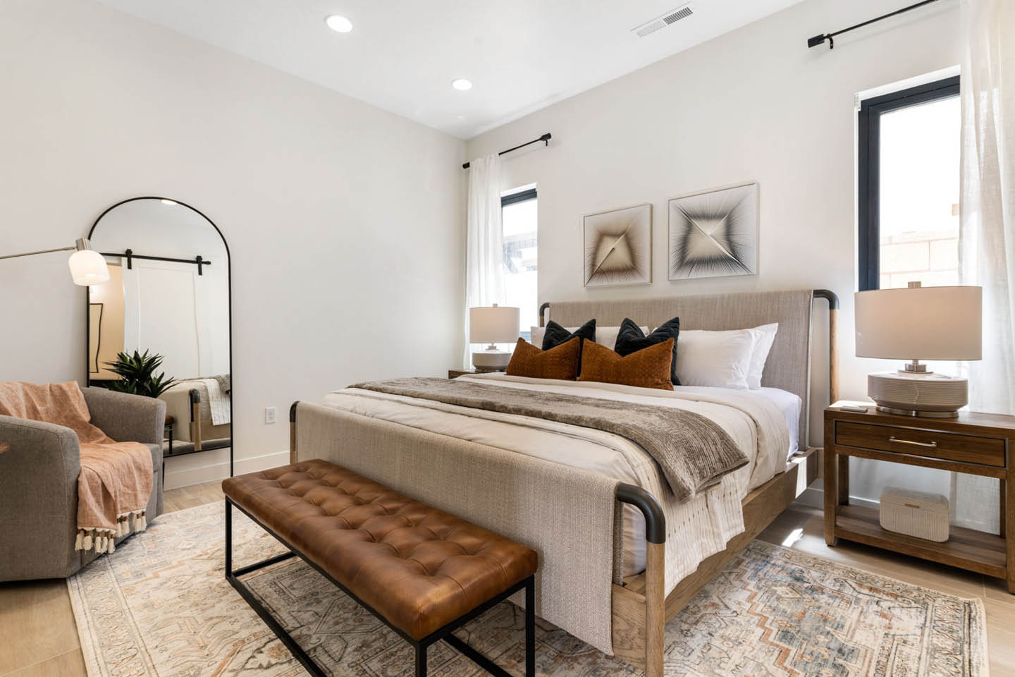 Primary bedroom with a king-size upholstered bed framed by walnut nightstands and ceramic table lamps, accented with black and rust-colored throw pillows. A tufted camel leather bench sits at the foot of the bed, a full-length arched floor mirror leans aga