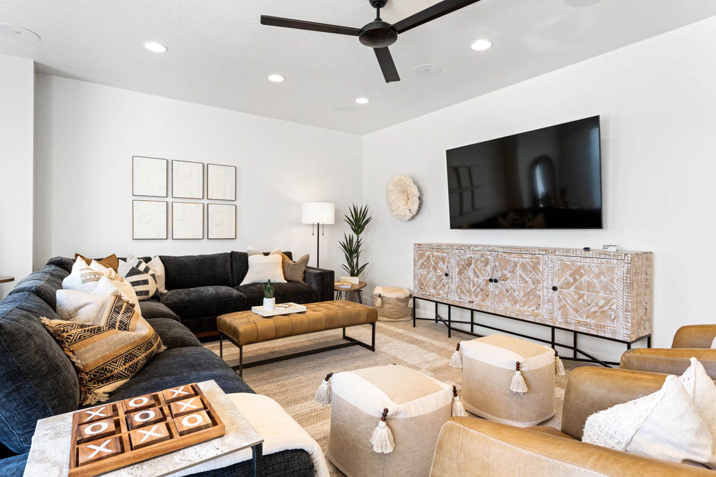 Media and family room with a large dark charcoal sectional sofa adorned with patterned bohemian-style pillows, a tufted tan leather ottoman, and a wall-mounted large-screen television above a carved whitewashed wood media console. Neutral poufs with tassel