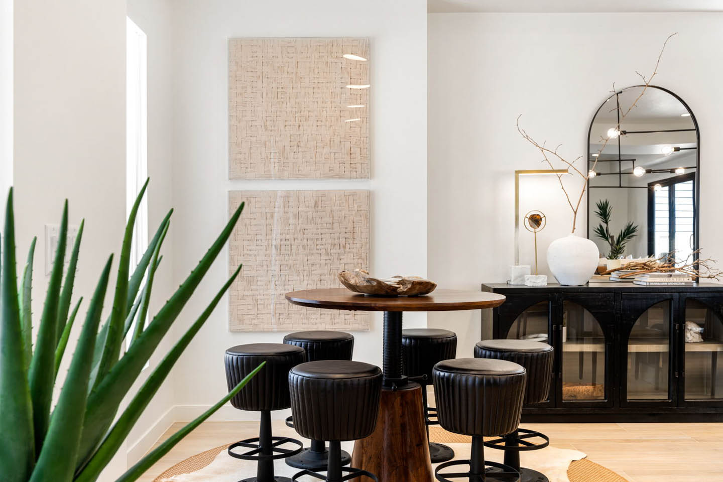 Casual dining nook with a round walnut pedestal table surrounded by black leather swivel stools, set on a natural jute rug beside a tall potted agave plant. Textured woven art panels hang on the wall above, while a black arched-door sideboard with an arche