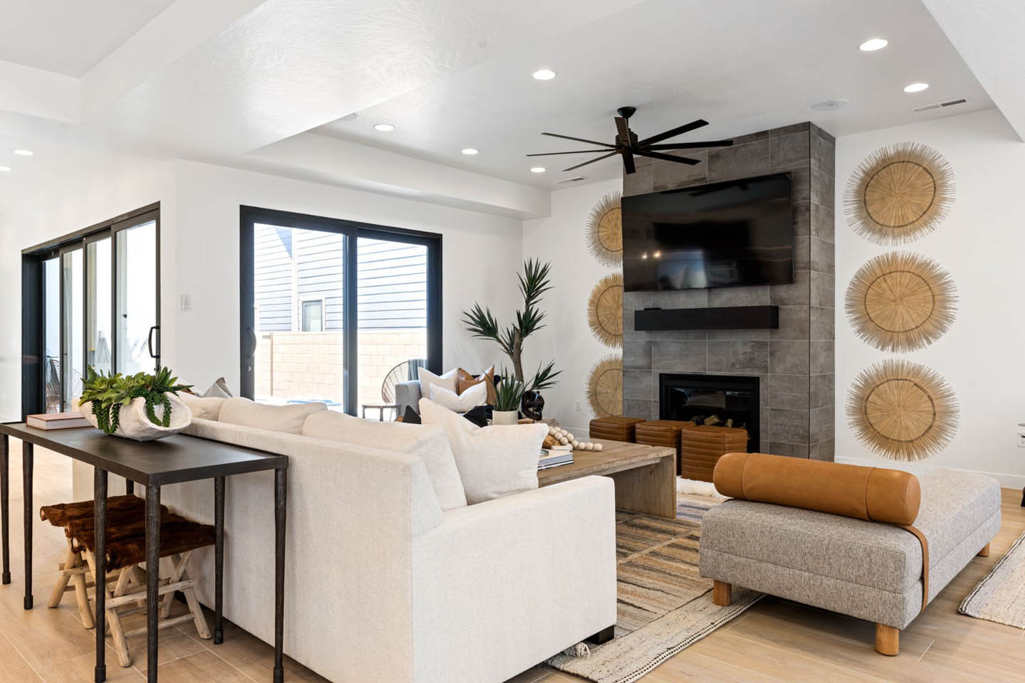 Living room with gray stone fireplace, wall-mounted TV, cream sectional, gray ottoman, woven wall accents, hardwood floors, ceiling fan, and black-framed sliding doors to a covered patio.