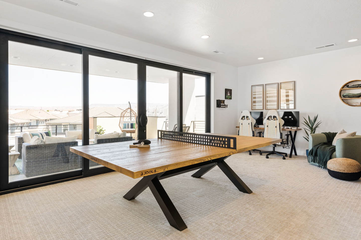 Recreation room featuring a designer wood-top ping pong table with matte black legs, a dual-monitor gaming station with two racing-style chairs, and a green sofa in the corner. Floor-to-ceiling black-framed sliding glass doors open to an upper-level balcon