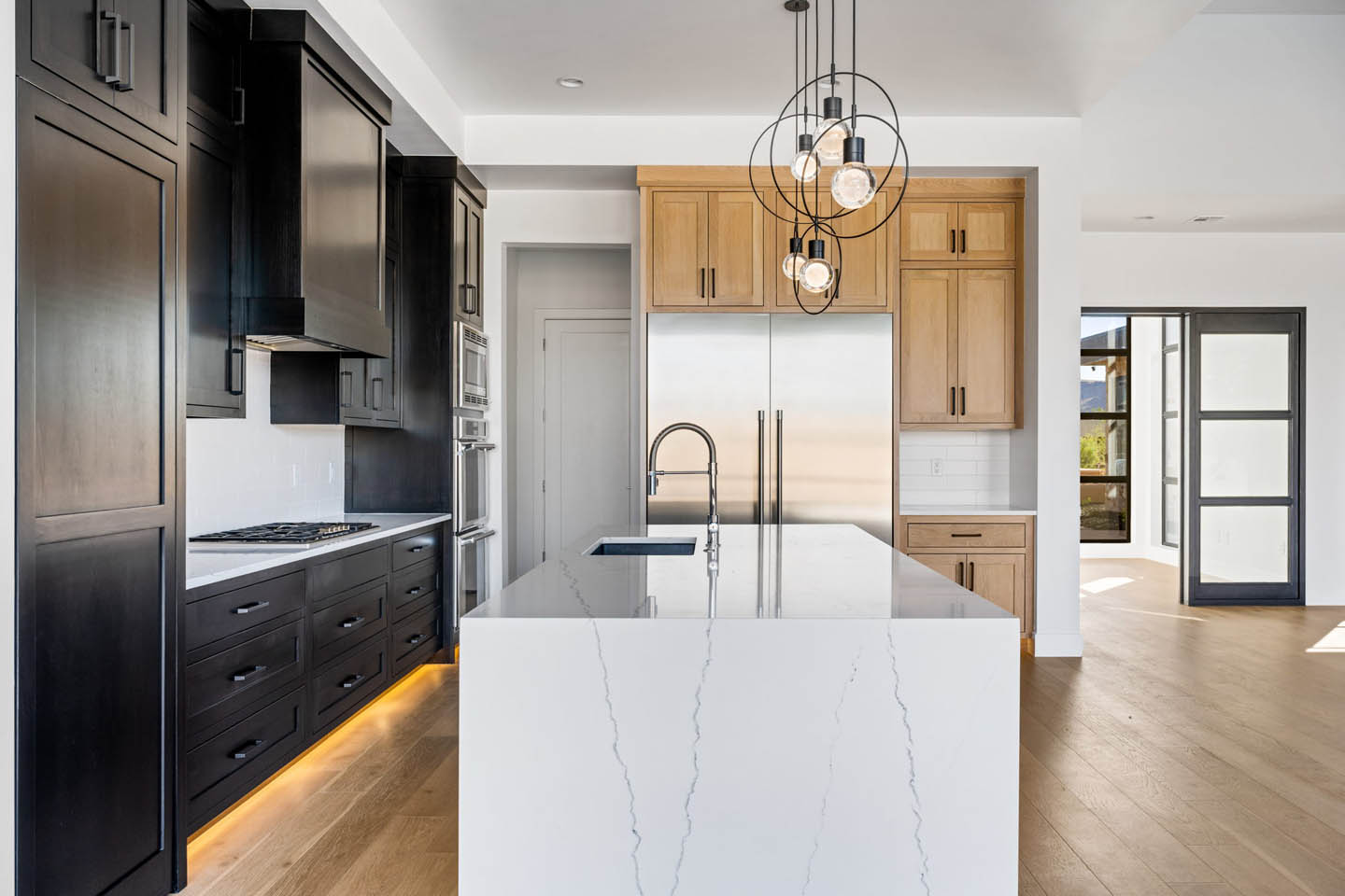 Modern kitchen with white marble waterfall island, dark floor-to-ceiling cabinetry with amber lighting, oak upper cabinets, built-in refrigerator, sculptural pendant lights, and wide-plank oak flooring.