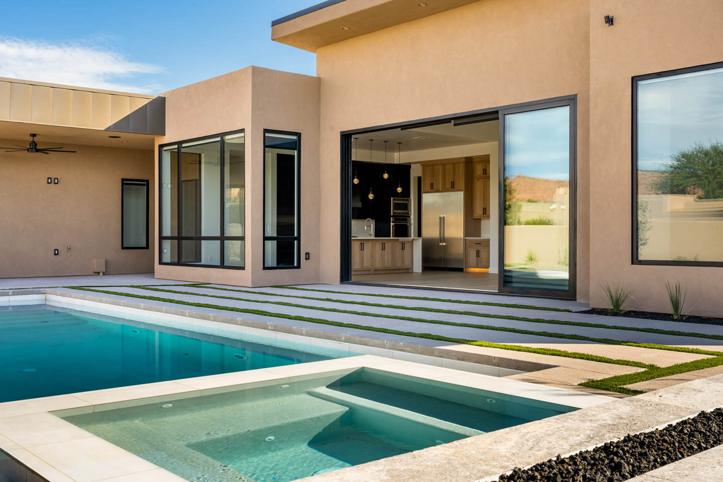 Contemporary desert home exterior with warm sand stucco, turquoise pool and spa, concrete paver and turf path, large retractable glass wall opening to the kitchen, and mountain landscape in the windows.