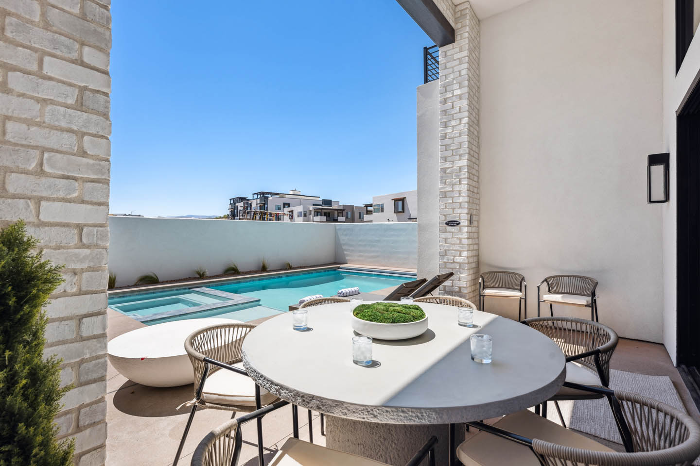 Covered patio dining area with round table and woven chairs beneath white stucco walls and stone columns, overlooking a turquoise pool and spa under a clear blue sky.