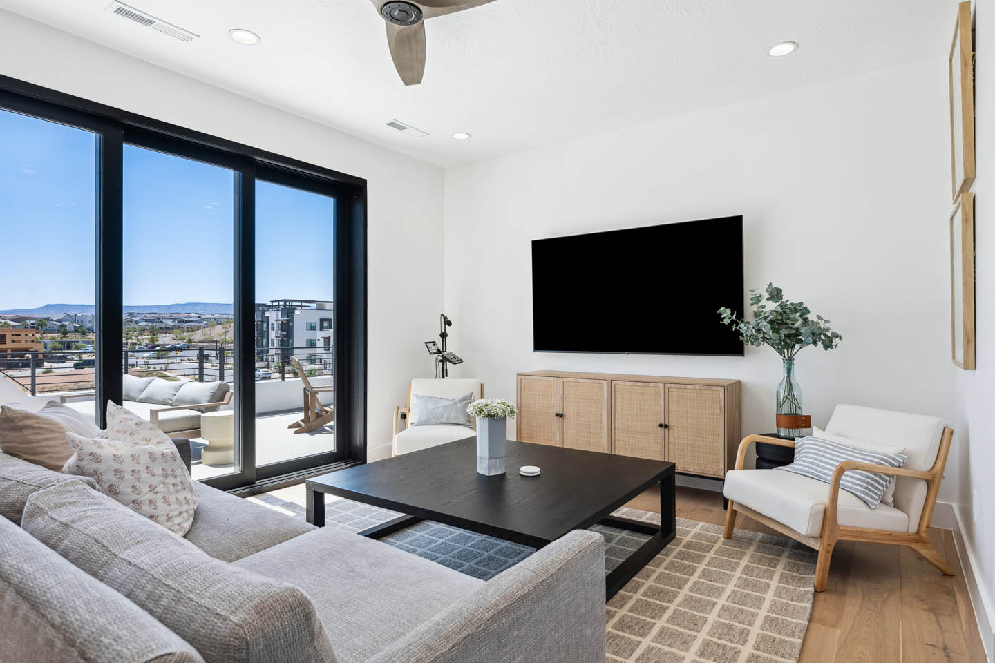 Upper-level living room with gray sectional, dark wood coffee table, rattan media console, wall-mounted TV, ceiling fan, and sliding glass doors opening to a balcony with mountain views.