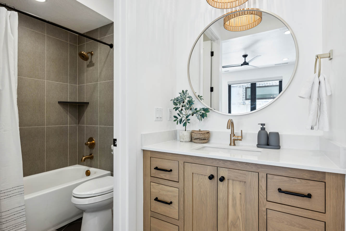 Guest bathroom with oak vanity, white quartz, brushed gold faucet, oval mirror, rattan pendant, and shower-tub combo with large-format taupe tile and brushed gold fixtures.