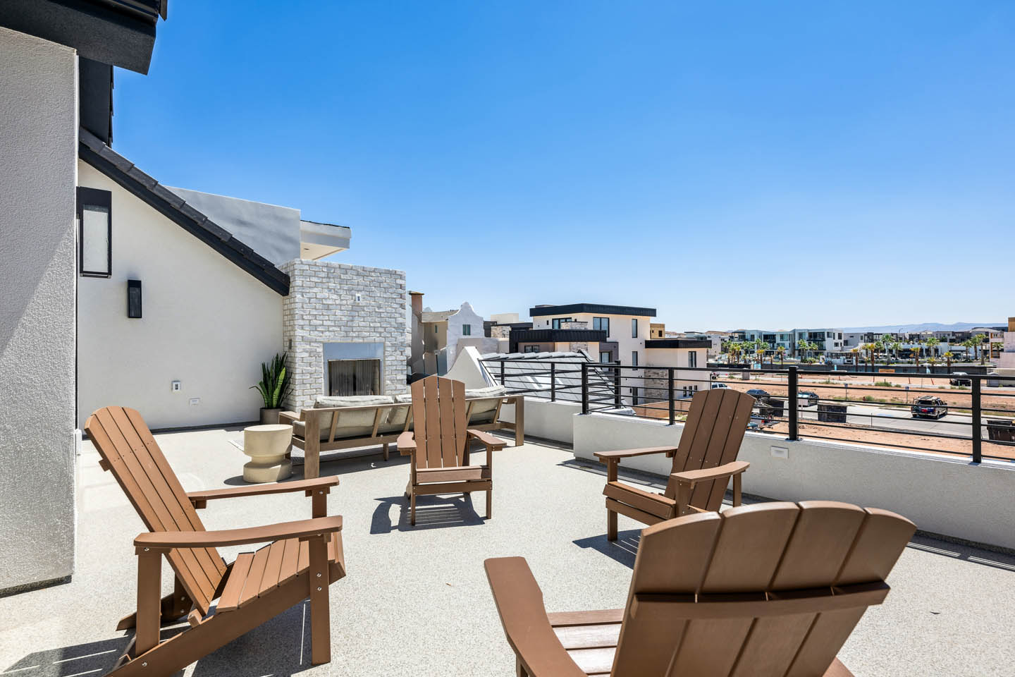 Rooftop terrace with brown Adirondack chairs around a stone fireplace, horizontal metal railing, and panoramic views of the surrounding neighborhood under a clear blue sky.