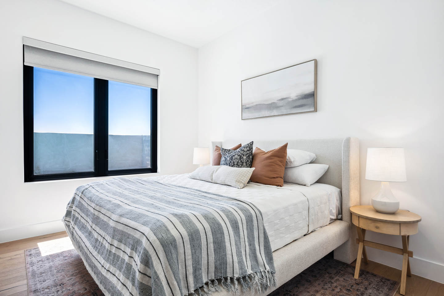 Guest bedroom with neutral upholstered bed, striped linen bedding, cognac accent pillows, round wood nightstand, ceramic table lamp, abstract wall art, and black-framed window.