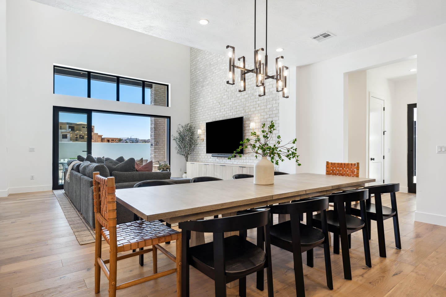 Open-concept living and dining area with double-height ceilings, wood dining table, black and woven leather chairs, modern chandelier, and white brick accent wall with mounted TV.