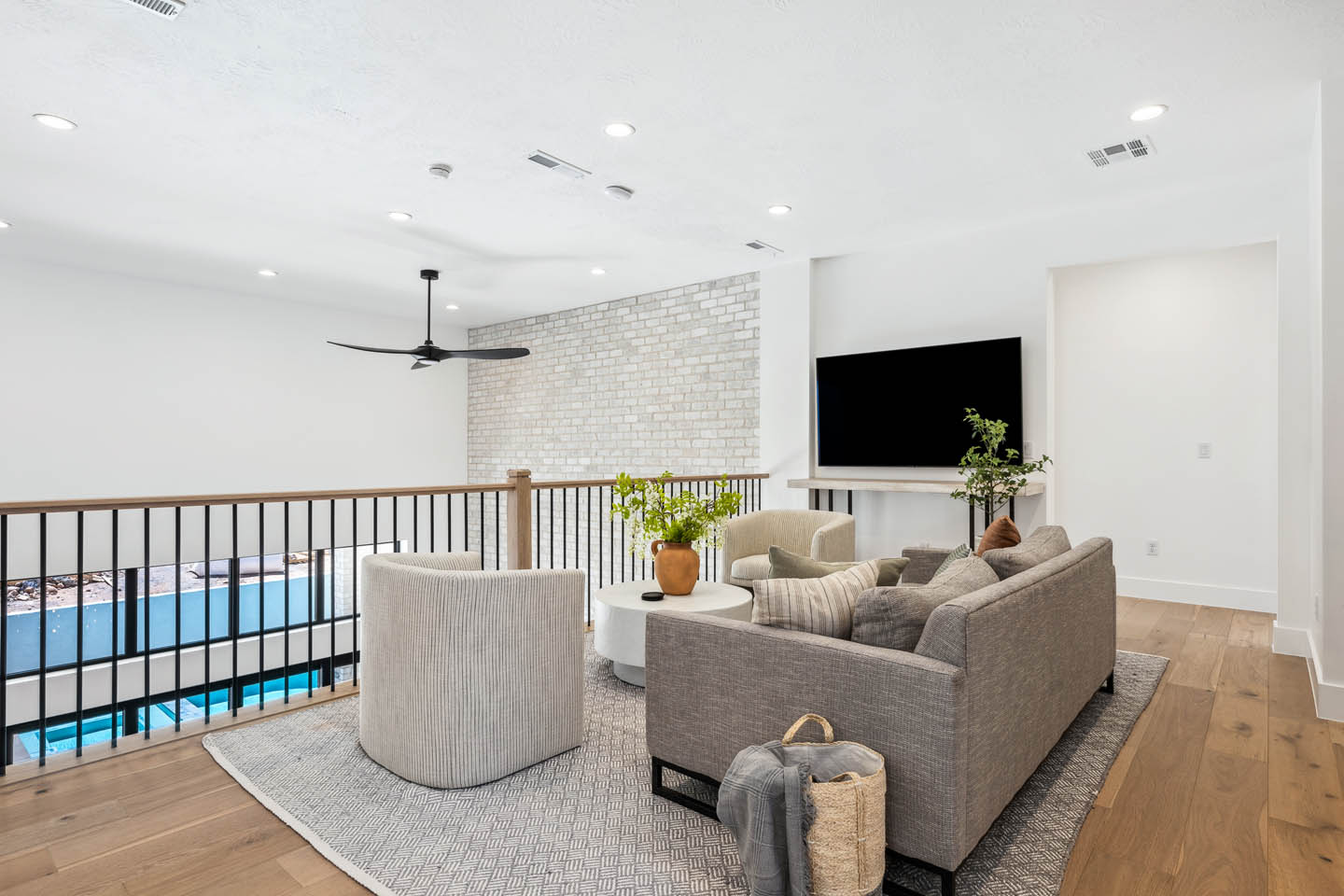Upper-level loft lounge with gray sofa and swivel chair, white side table, textured rug, wall-mounted TV on white brick, black ceiling fan, and metal-and-wood pool-view railing.