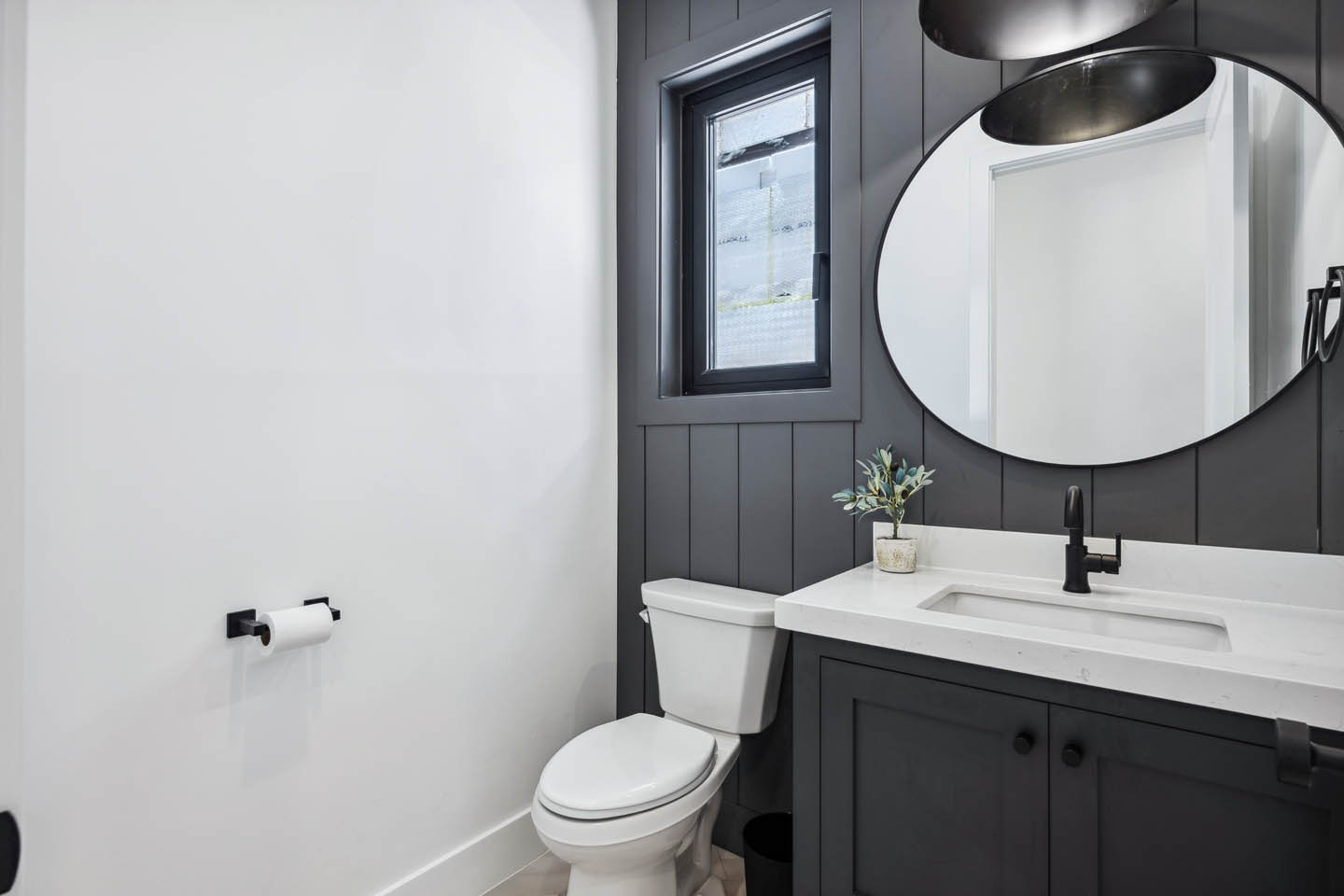 Powder room with dark charcoal vertical shiplap wall, dark vanity, white countertop, matte black faucet, large round mirror, dome pendant light, and black-framed privacy window.