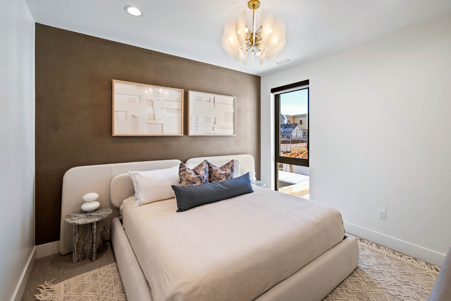 Guest bedroom with upholstered bed, taupe accent wall, floral and navy pillows, paired abstract artwork, marble-topped side table, and a brass and glass chandelier overhead.