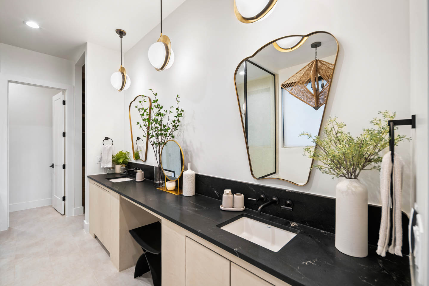 Bathroom vanity with dark marble countertop, light wood cabinetry, dual undermount sinks, matte black wall-mounted faucets, organic gold-framed mirror, and sculptural gold pendant lights.