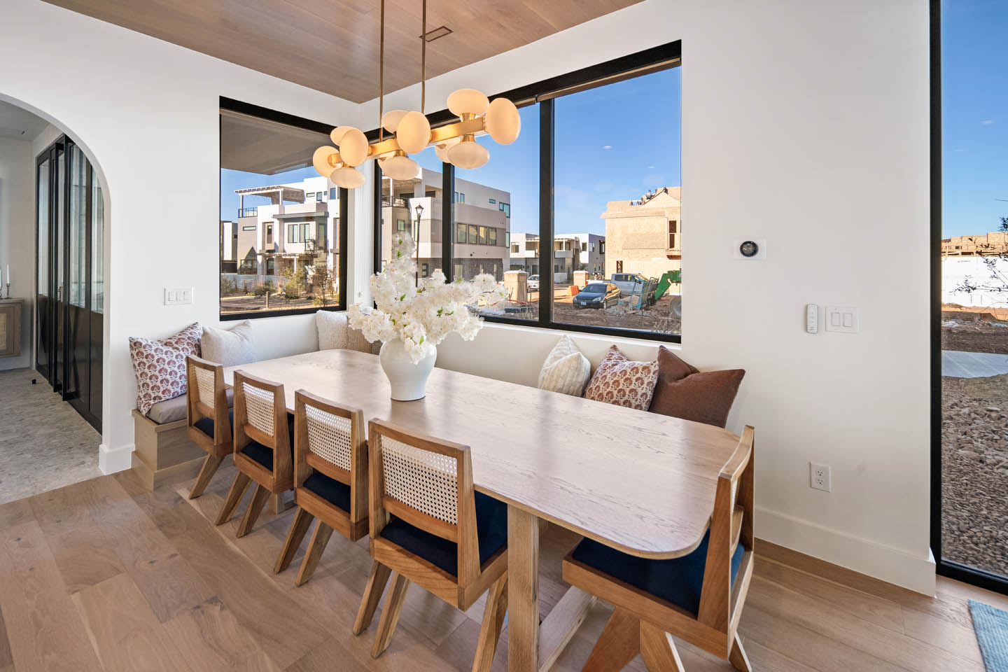 Dining nook with light wood table, cane-back chairs, built-in banquette, gold globe chandelier, wood-paneled ceiling, and black-framed windows overlooking the surrounding community.