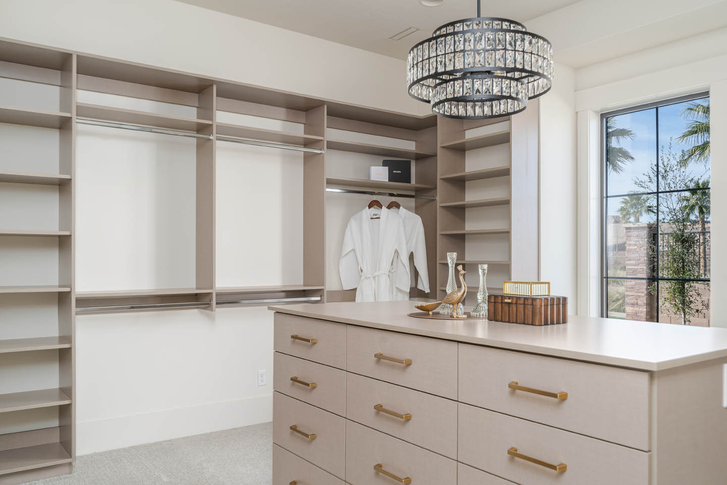 Walk-in closet with floor-to-ceiling custom built-ins in warm taupe, dresser island with brushed gold pulls, crystal chandelier, and black-framed window with palm views.