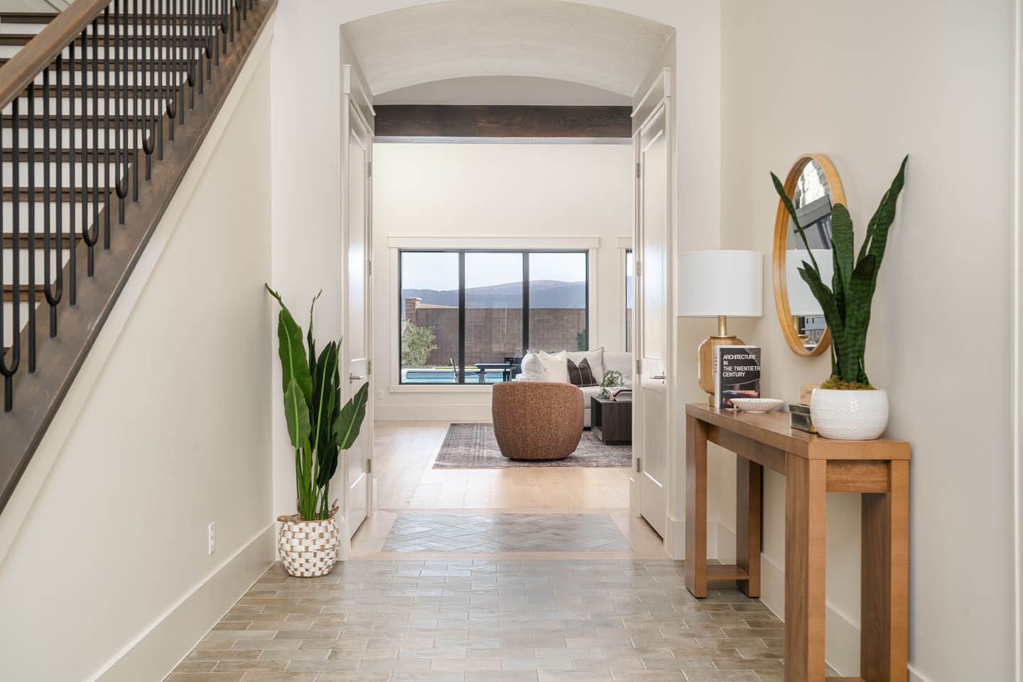 Entry hallway with herringbone tile floors, wood console, round mirror, snake plant, and a staircase with dark treads and black metal balusters leading to the upper level.