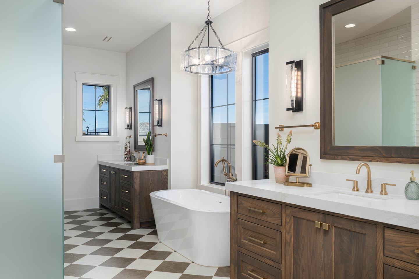 Primary bathroom with dual dark wood vanities, white quartz, brushed gold fixtures, freestanding soaking tub, checkered floor tile, and a glass pendant chandelier.