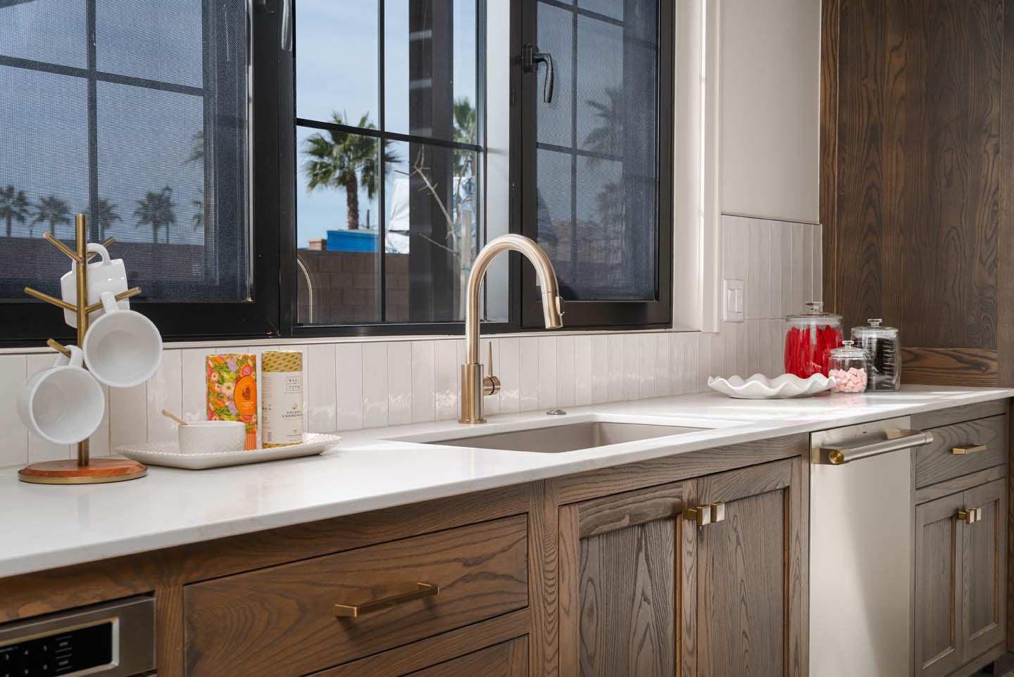 Kitchen prep area with white quartz countertops, vertical white tile backsplash, dark oak cabinetry with brushed gold hardware, and a gooseneck faucet below casement windows.