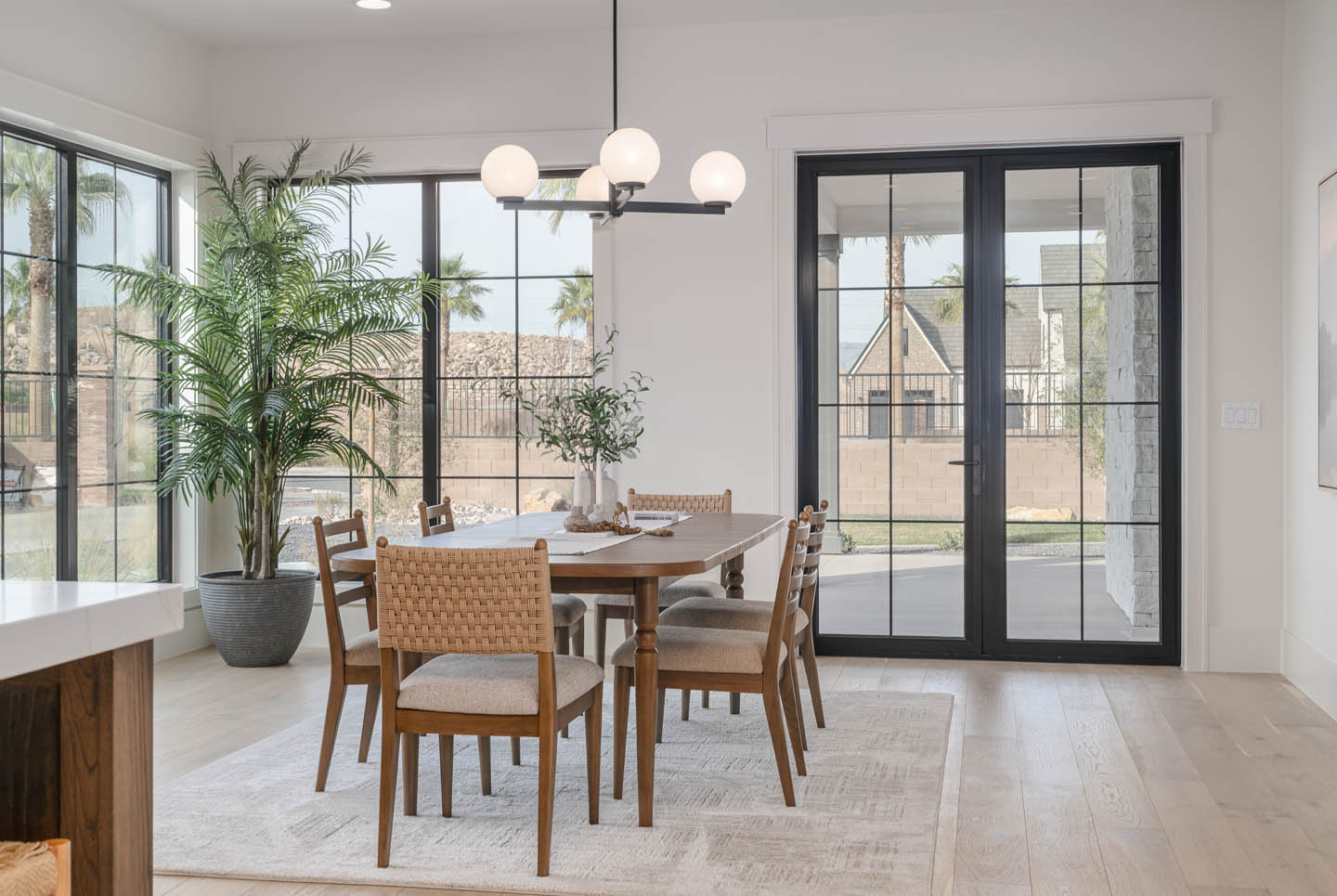 Dining area with mid-century wood table, woven chairs, globe chandelier, and floor-to-ceiling black-framed steel windows flooding the space with natural light.