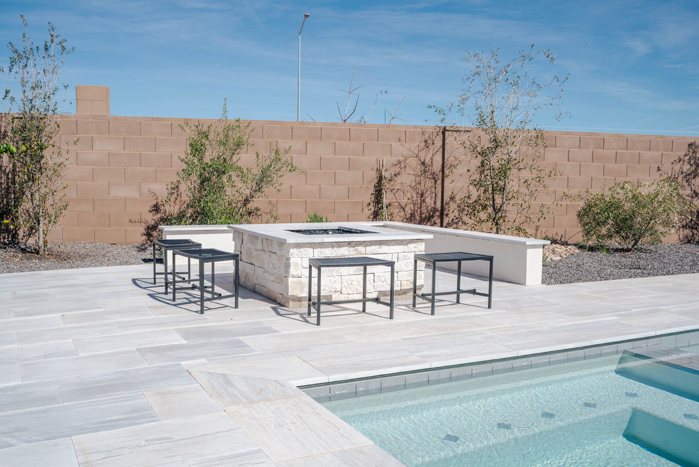 Poolside entertaining area with L-shaped stone fire pit bar, black metal counter stools, turquoise swimming pool, and desert landscaping under an open blue sky.
