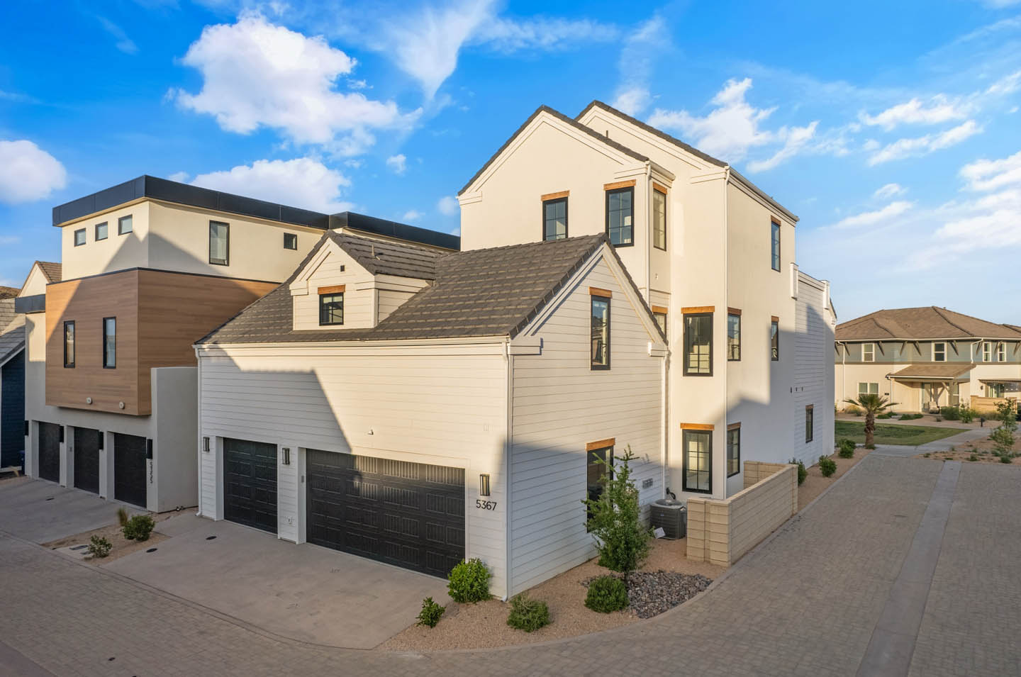 Three-story home exterior with white lap siding, cream stucco upper levels, wood accent trim, black-framed windows, dark garage doors, and desert landscaping.