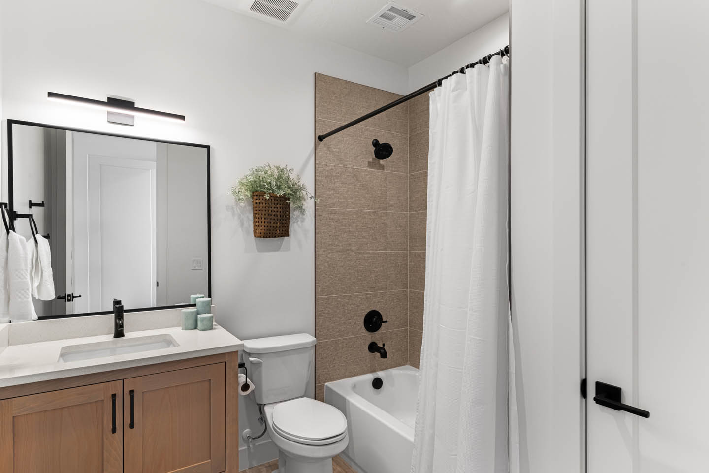 Guest bathroom with warm wood vanity, white quartz countertop, black-framed mirror, matte black fixtures, tub-shower combo with tan tile, and a tall white linen cabinet.