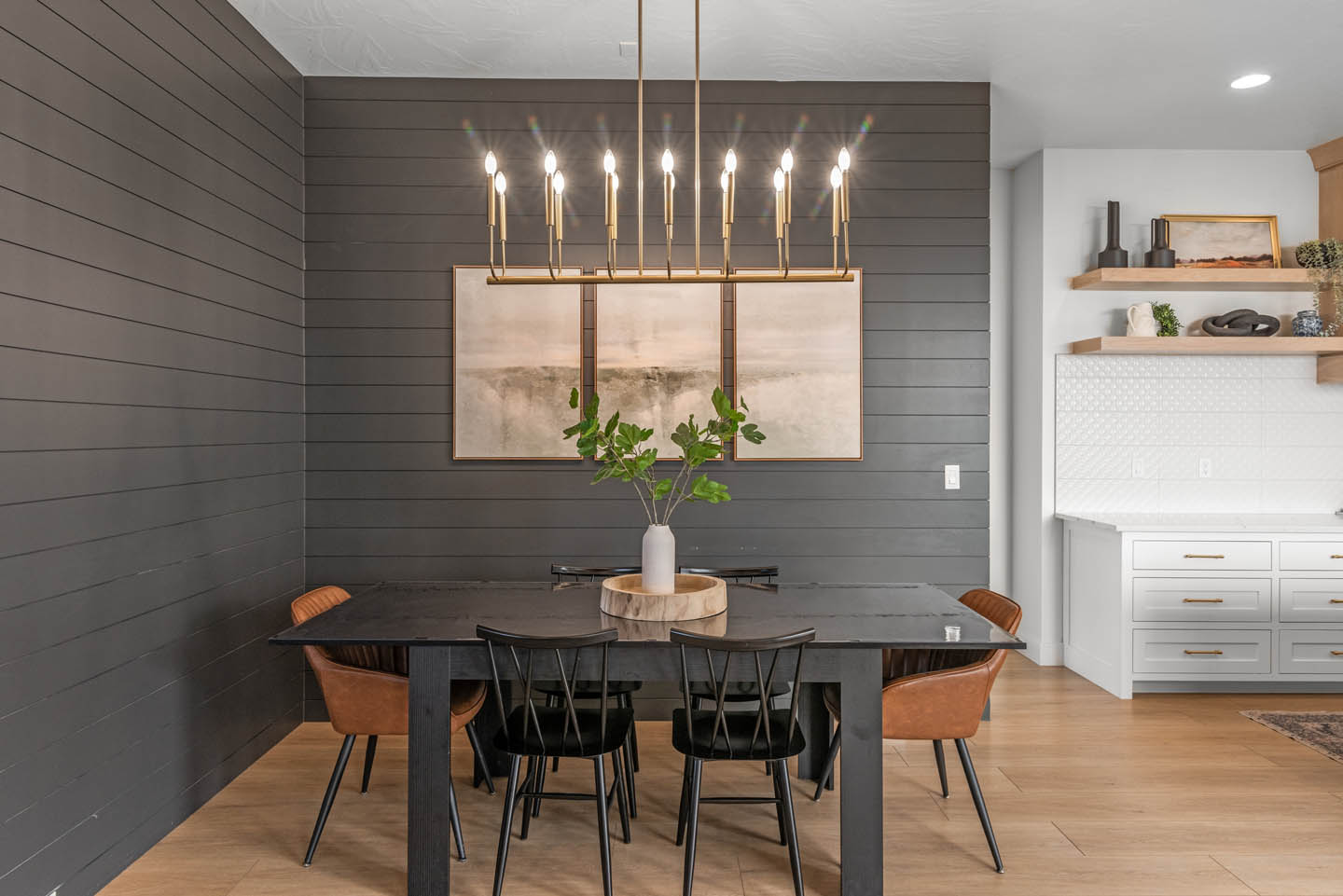 Dining area with dark charcoal shiplap walls, black table, spindle-back and cognac leather chairs, gold linear chandelier, and white kitchen with floating shelves visible beyond.
