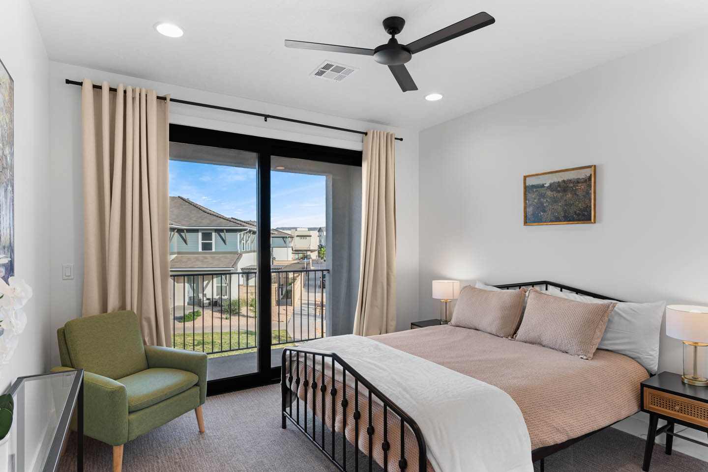Secondary bedroom with white walls, black metal-frame bed, blush and white bedding, green accent chair, and black-framed sliding glass doors opening to a balcony.