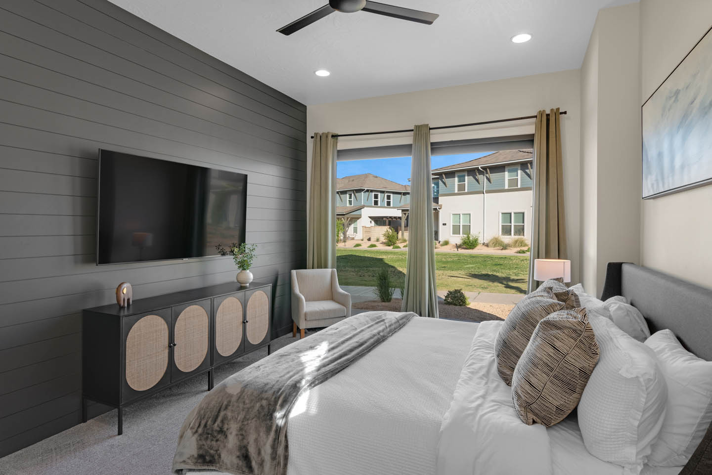 Bedroom with dark gray shiplap accent wall, wall-mounted TV, black-and-cane media console, king bed in white and gray bedding, gold drapes, and a black ceiling fan.