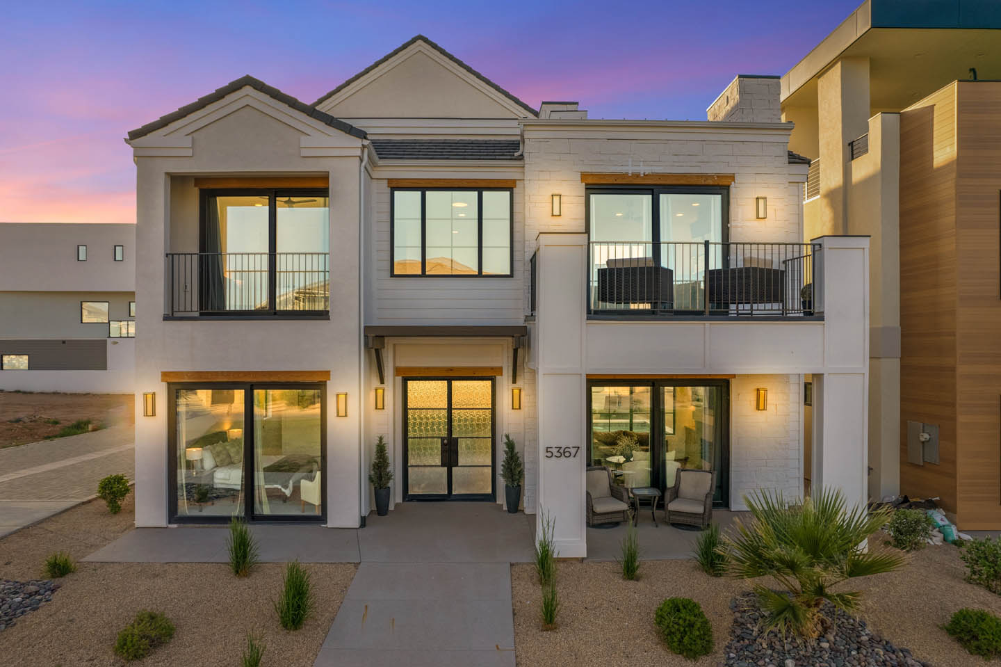 Twilight exterior of a two-story white stucco and stone home with black-framed windows, iron balcony railings, wood beam accents, desert landscaping, and a vivid sunset sky.