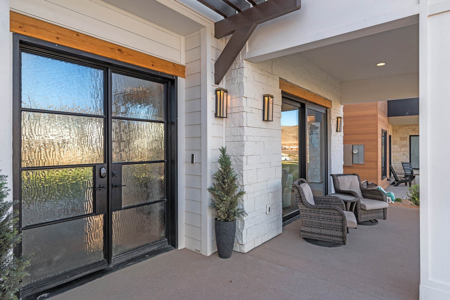 Covered front entry porch with black-framed French doors, white stone columns, dark wood beam brackets, wicker lounge chairs, black wall sconces, and potted greenery.
