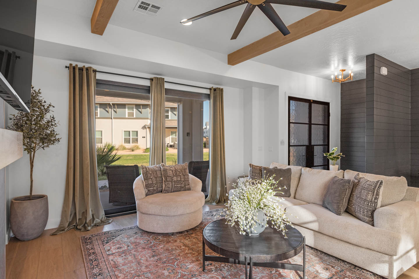 Living room with vaulted ceilings, exposed wood beams, ceiling fan, neutral linen sectional, Persian rug, dark wood coffee table, gray shiplap wall, and sliding glass doors.