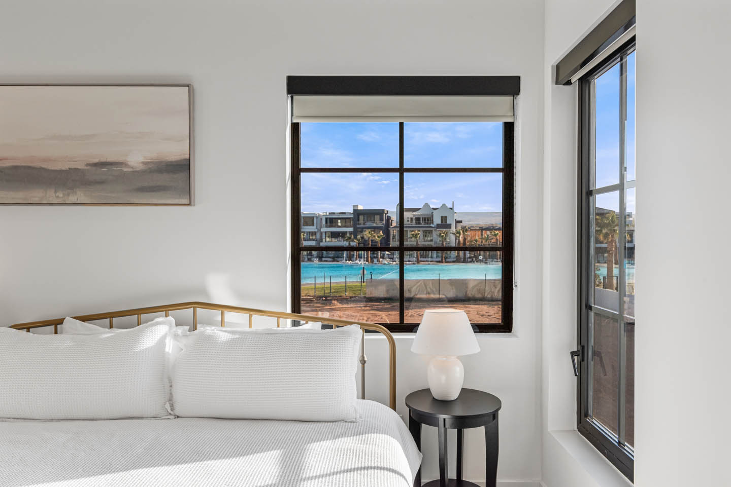 Bedroom with gold metal-frame bed, white waffle-textured bedding, round black nightstand, ceramic lamp, and black-framed grid windows overlooking a turquoise community lagoon.
