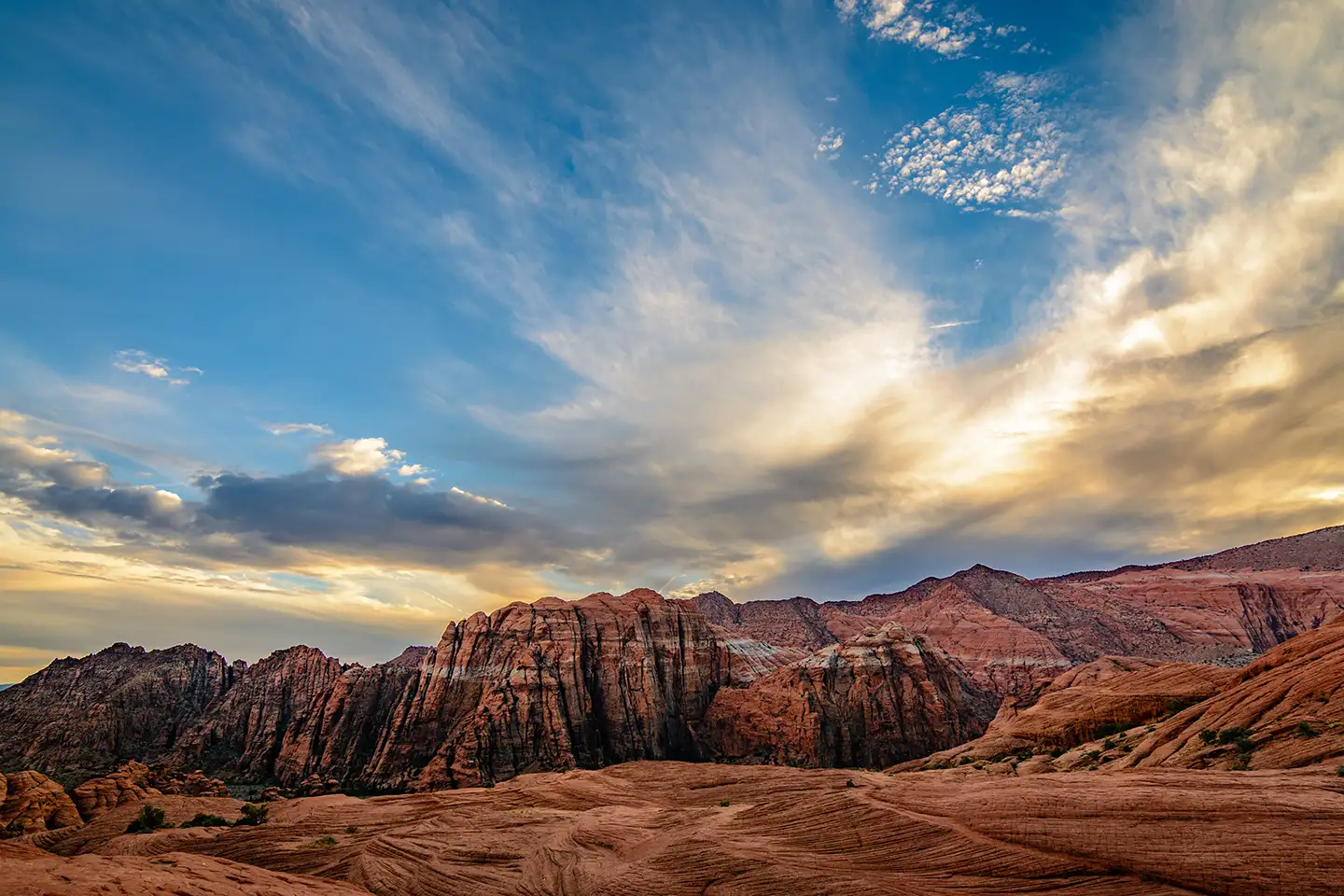 Dramatic layered red sandstone cliffs and sweeping petrified sand dunes beneath a golden-blue sunset sky in the Utah desert.