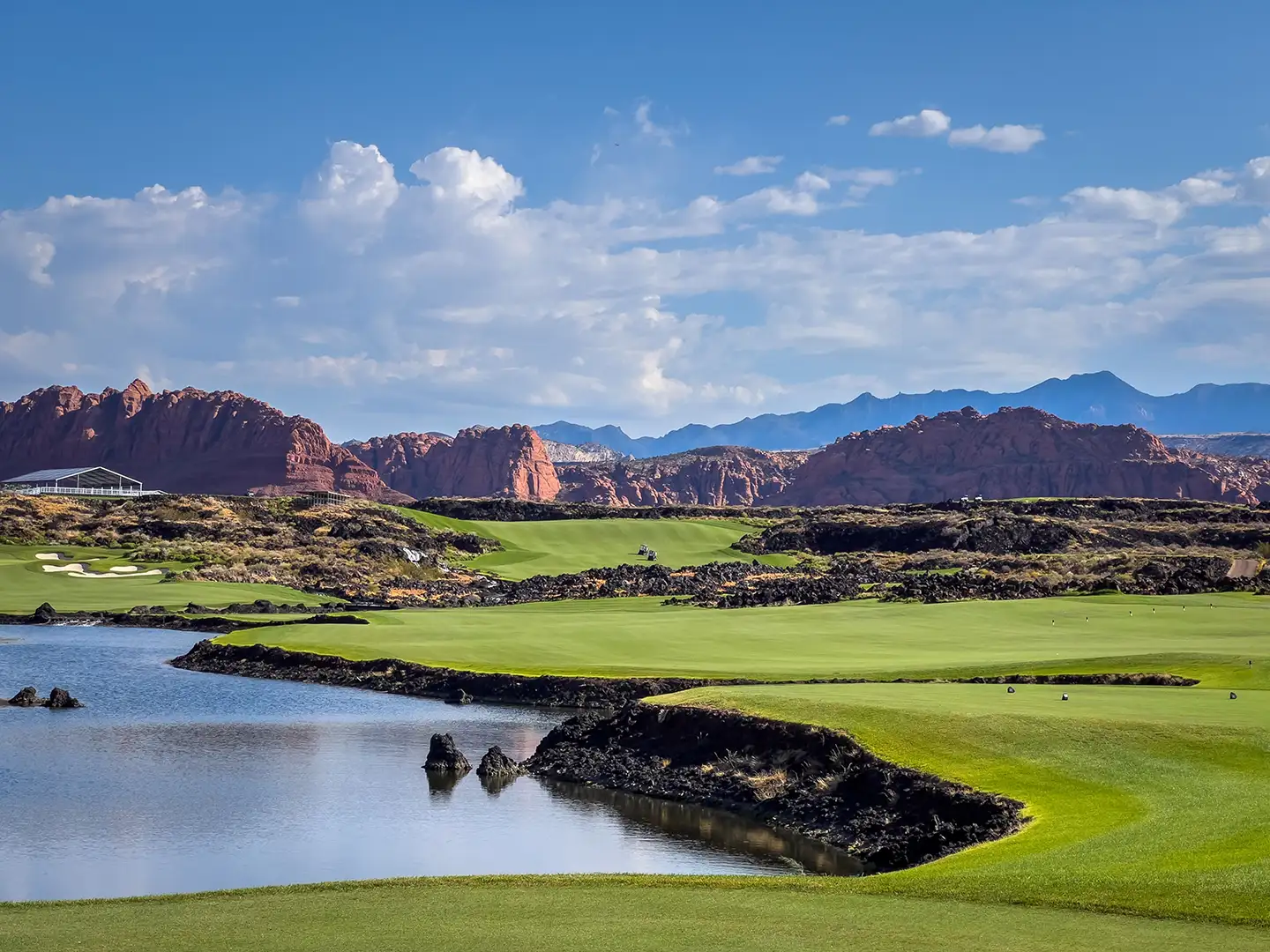 Lush green golf course hole with a water hazard edged by black lava rock, framed by dramatic red sandstone formations under a blue sky.