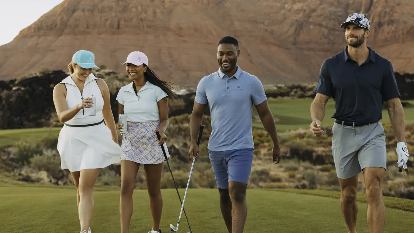 Four smiling golfers in colorful attire walking a sunlit fairway with towering red rock desert cliffs rising behind them.