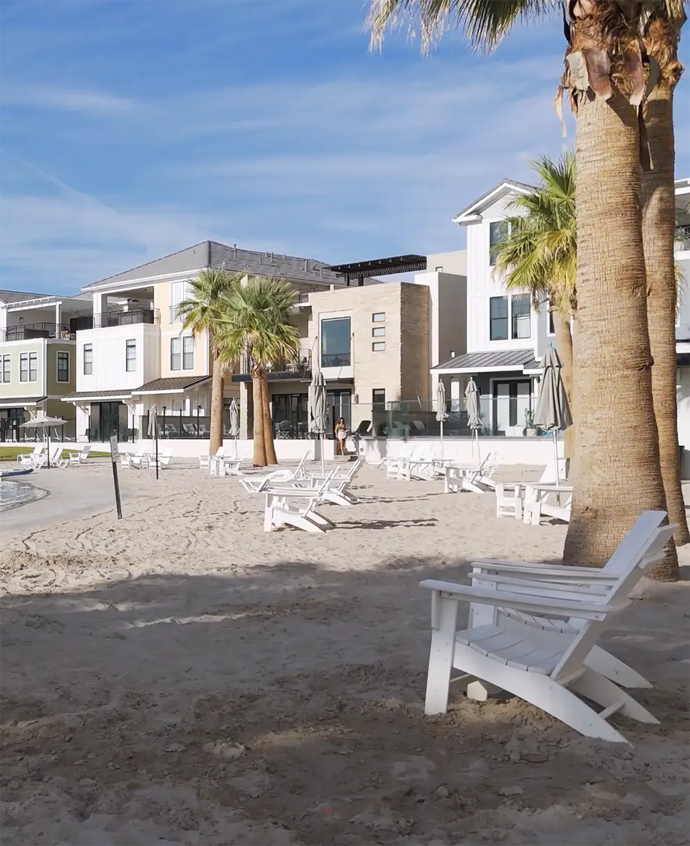 White Adirondack chairs on a sandy beach with palm trees and modern Desert Color townhomes under a clear blue sky.