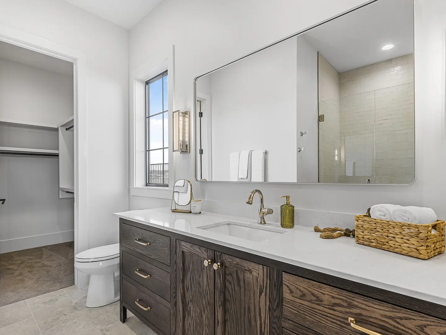 Primary bathroom featuring a dark oak vanity with quartz countertop, gold fixtures, large mirror, and walk-in closet at Cedar Pointe Homes.
